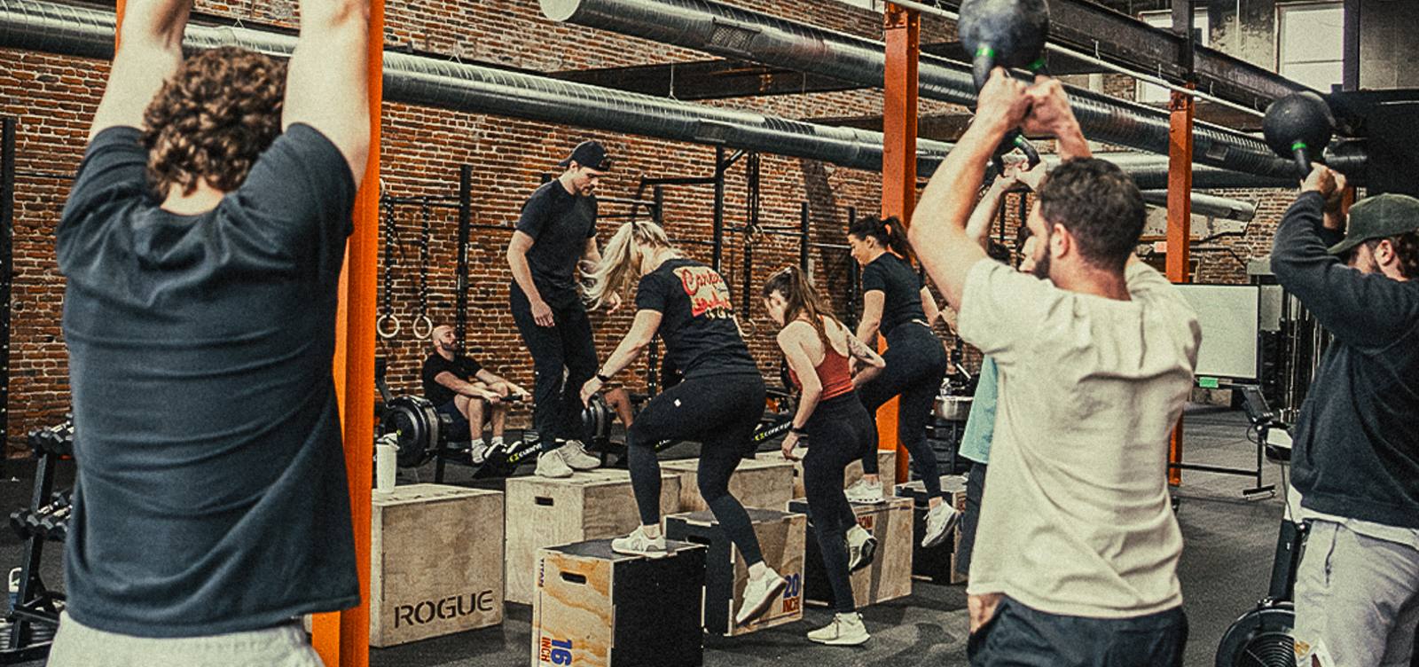 A packed group class at Omen Athletics: kettlebell swings in front, box step-ups and rowers behind, orange steel columns and brick walls.